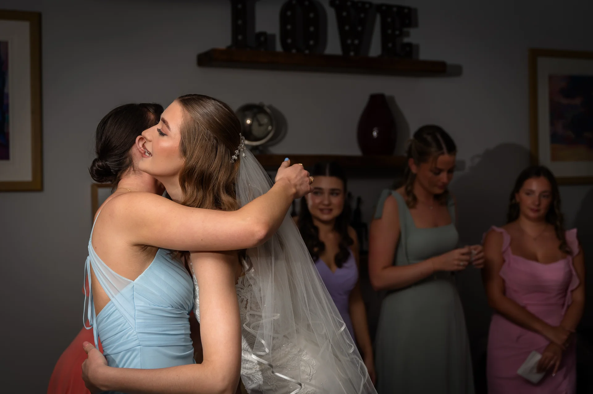 A smiling bride wearing a lace wedding dress and fingertip veil shares a warm embrace with a bridesmaid in a blue dress during morning preparations.