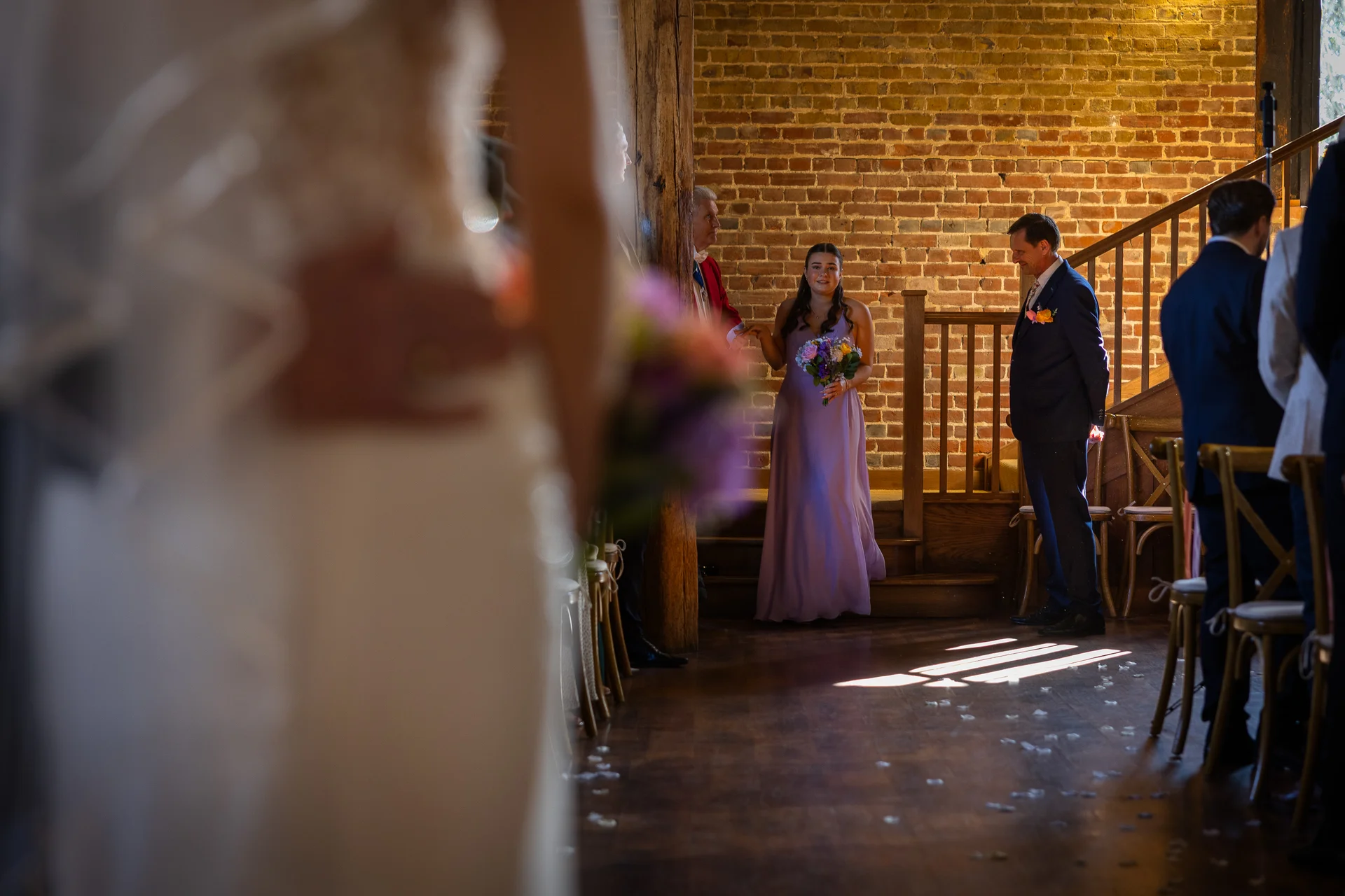 A bridesmaid in a lilac dress holding a bouquet waits at the top of the aisle inside a rustic barn with exposed brick walls.