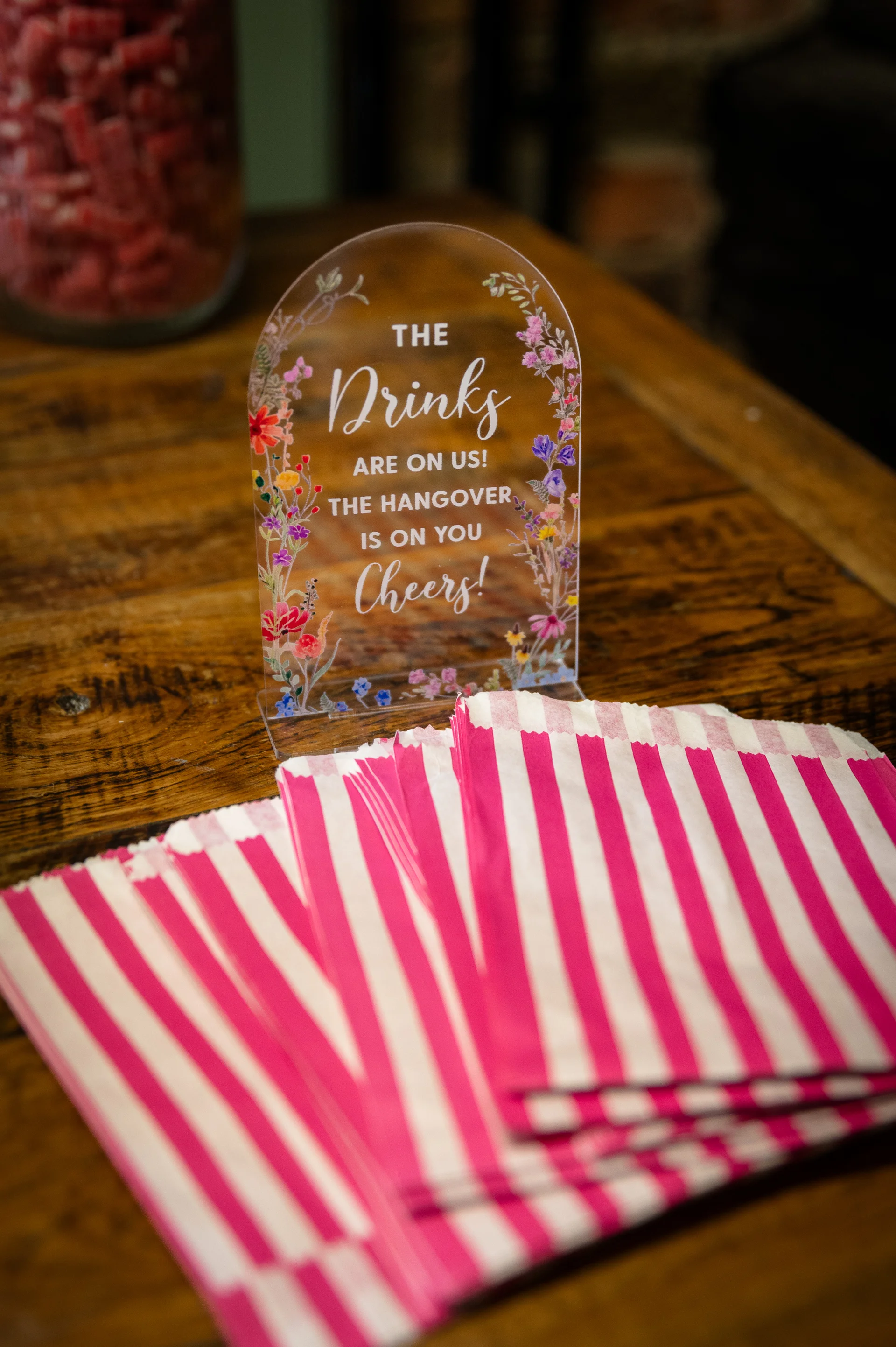 An arched acrylic sign with floral borders sits on a wooden table beside a stack of pink and white striped sweet bags.