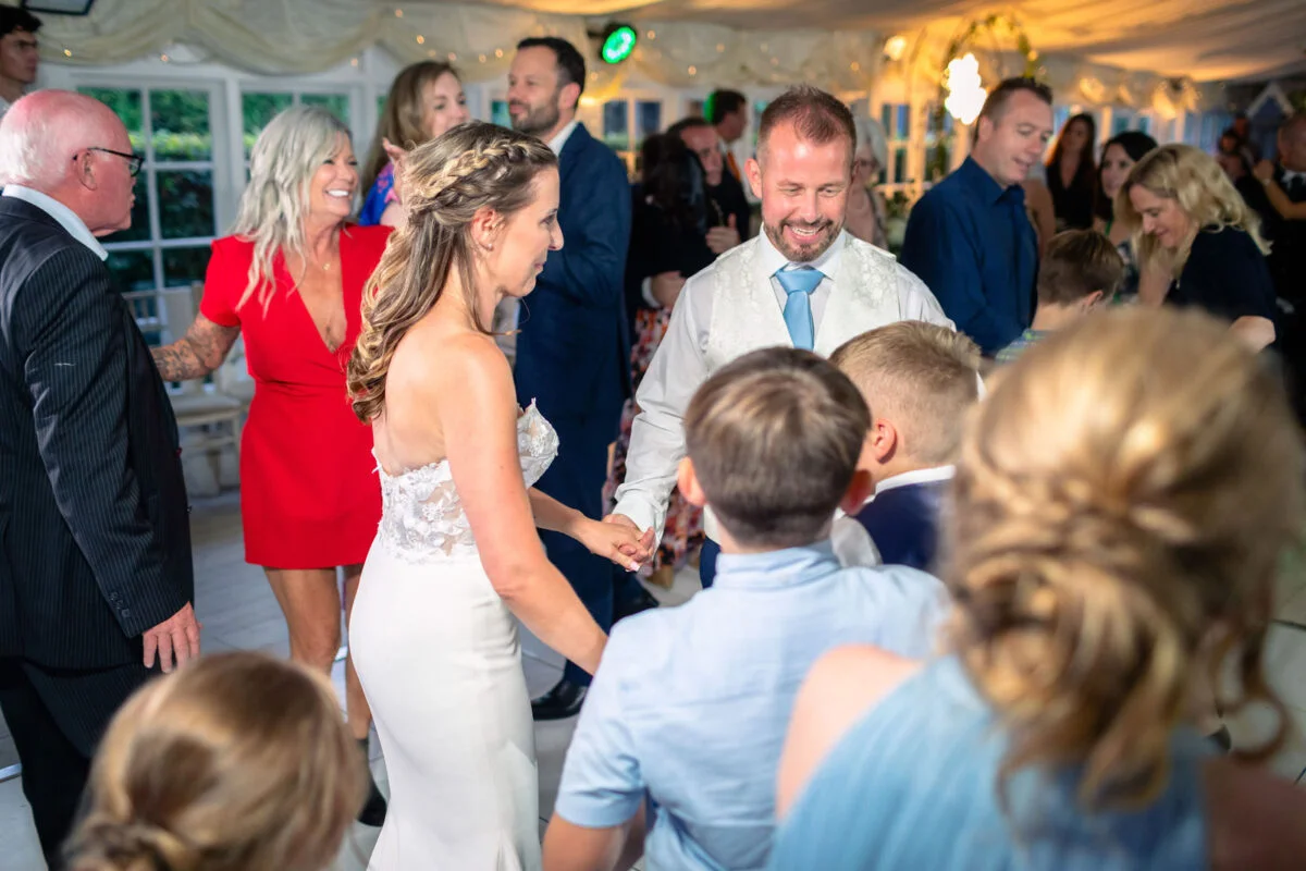 The bride and groom share a joyful first dance on the crowded dance floor surrounded by cheering family and guests inside a brightly lit marquee.