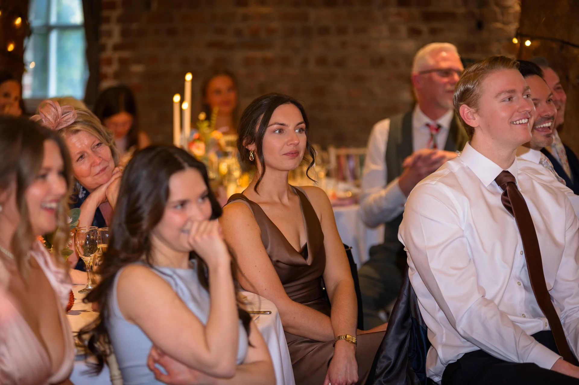 Wedding guests laugh and smile as they listen attentively to speeches in an exposed brick reception venue.