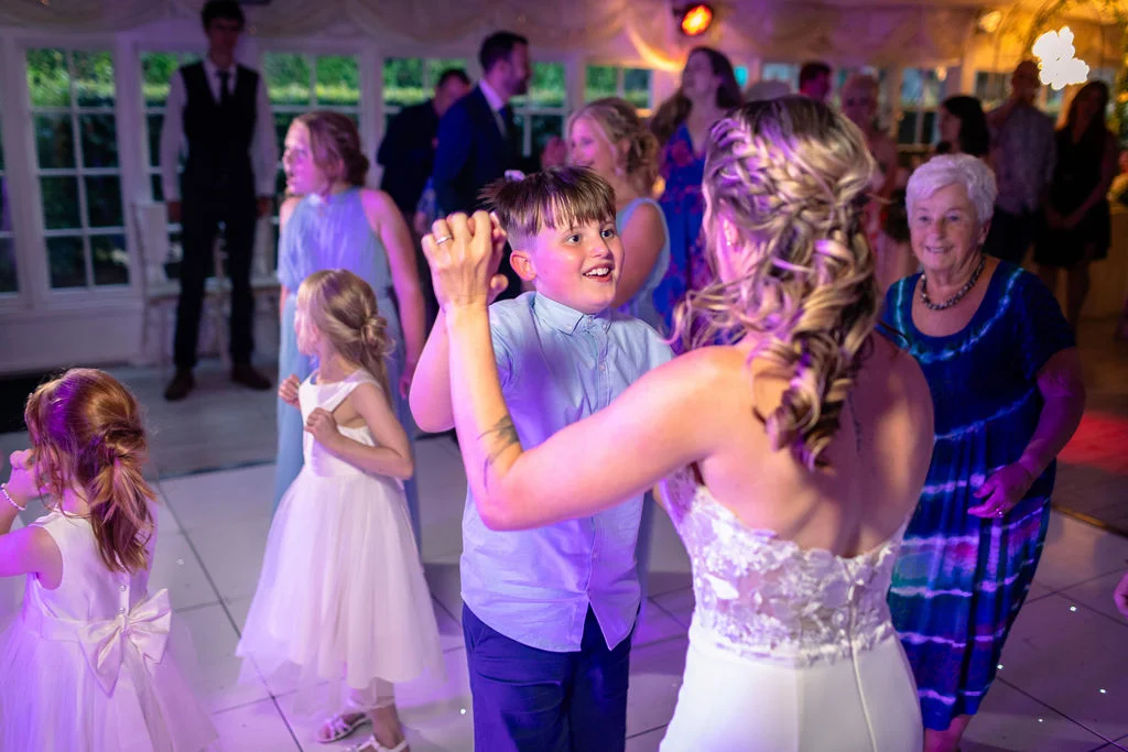 A young boy smiles joyfully as he dances with the bride on a lit-up dance floor surrounded by smiling wedding guests.