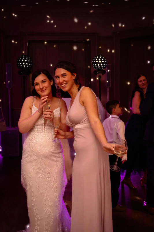 Two smiling brides holding champagne glasses pose together during an evening wedding reception illuminated by disco lights and mirror balls.