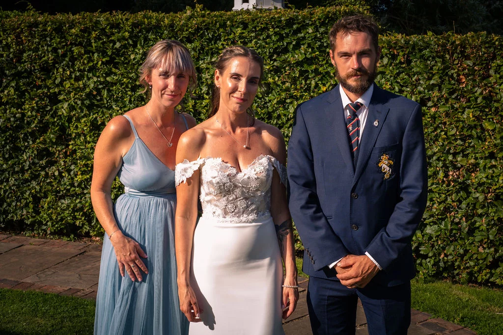 A bride in an off-the-shoulder lace dress stands alongside a bridesmaid in a dusty blue gown and a groom in a navy suit outdoors against a green hedge.