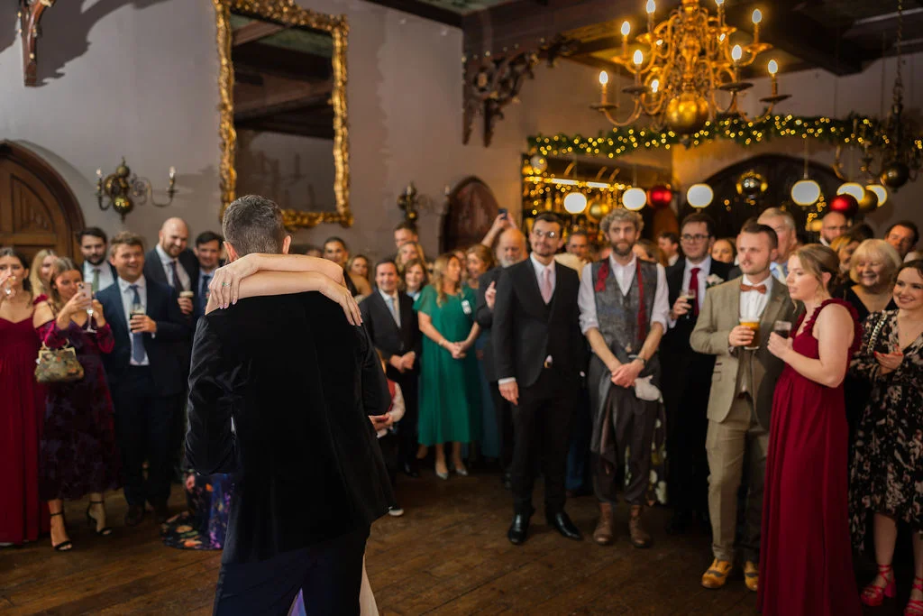 A newlywed couple shares an intimate embrace during their first dance, watched by a crowd of guests in a grand, chandelier-lit hall.