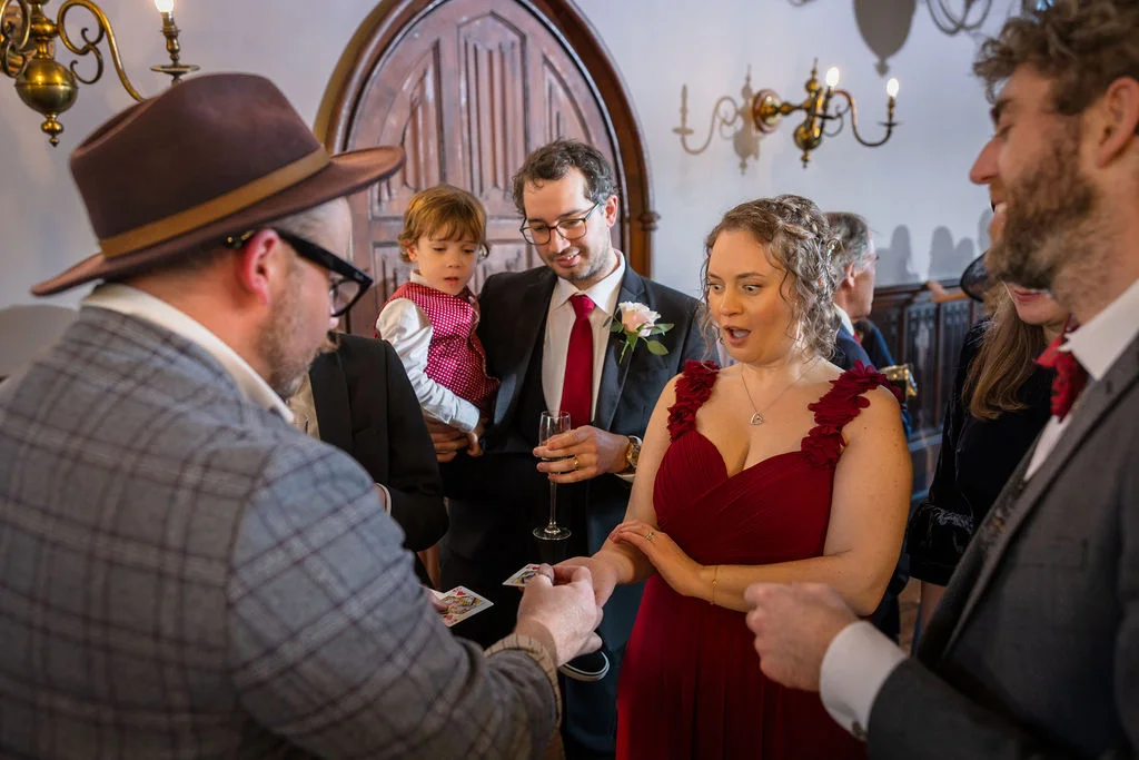 A wedding guest in a red dress looks on in astonishment as a close-up magician performs a card trick during the drinks reception.