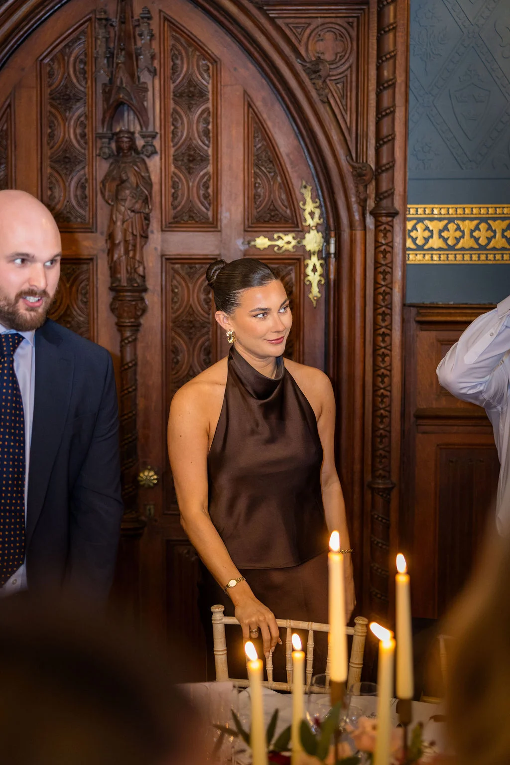 An elegantly dressed wedding guest in a brown halterneck top stands in a wood-panelled room next to a candlelit table.