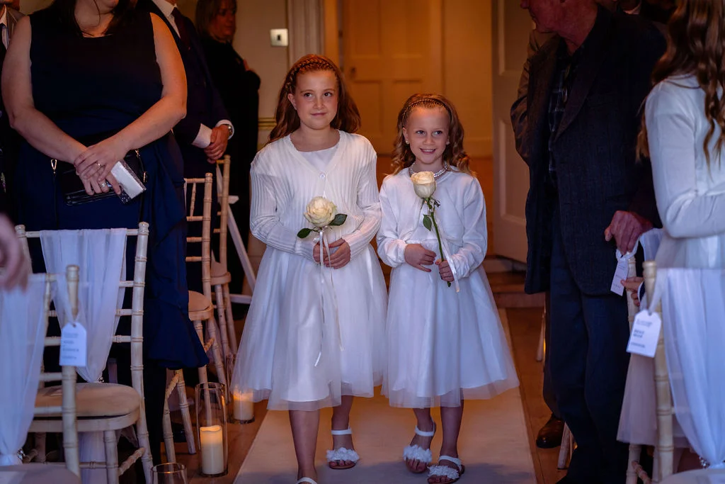 Two young flower girls in white dresses walk down the aisle holding single white roses during a wedding ceremony.