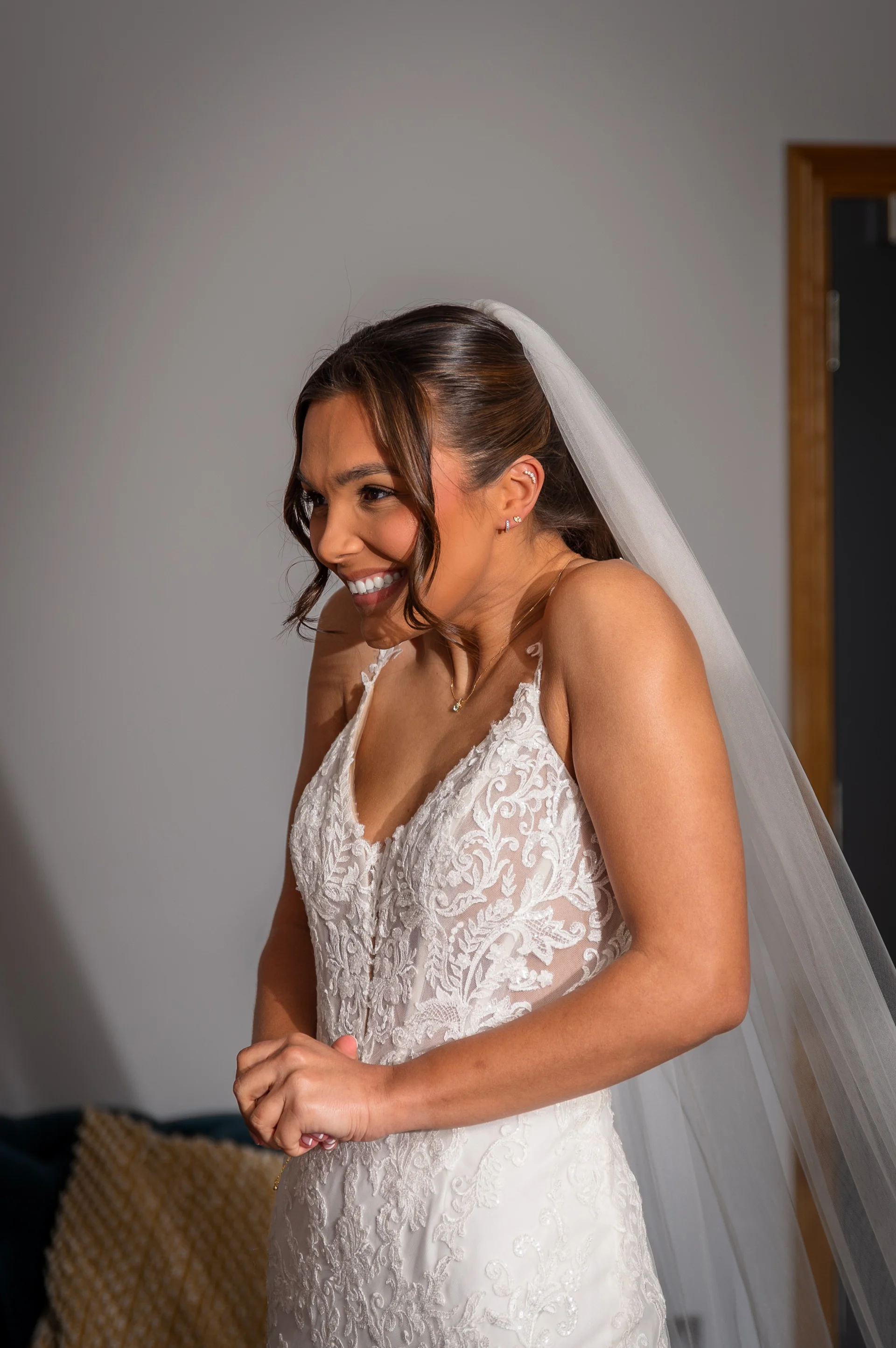 A smiling bride with half-up hair and face-framing tendrils wears a lace v-neck wedding dress and veil while getting ready.
