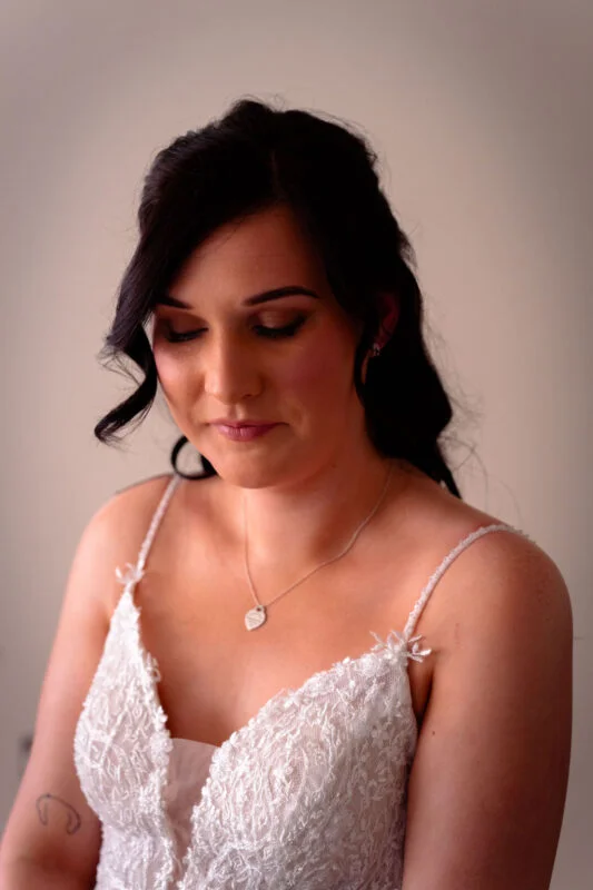 A close-up portrait of a dark-haired bride with half-up wavy hair, wearing a lace dress with spaghetti straps and a delicate necklace.