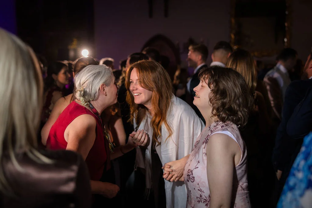 Wedding guests laugh and socialise together on a crowded evening dance floor under warm event lighting.