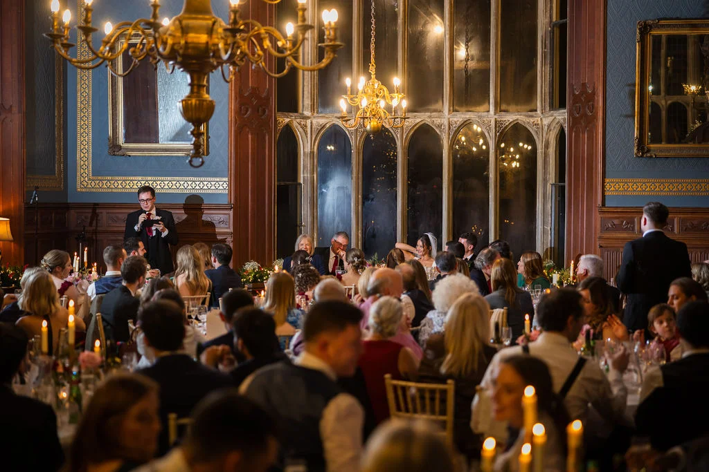 A best man delivers a speech during a wedding breakfast in a grand, wood-panelled stately home dining room illuminated by chandeliers and candlelight.