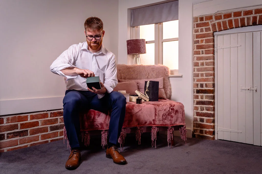 A groom in a white shirt and trousers sits on a pink-draped sofa indoors, opening a small gift box as natural light streams through the window.