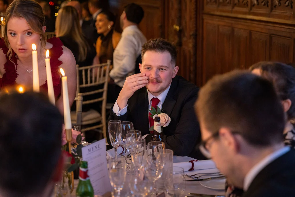 A guest sits at a candlelit wedding reception table listening to speeches while other guests chat in the background.