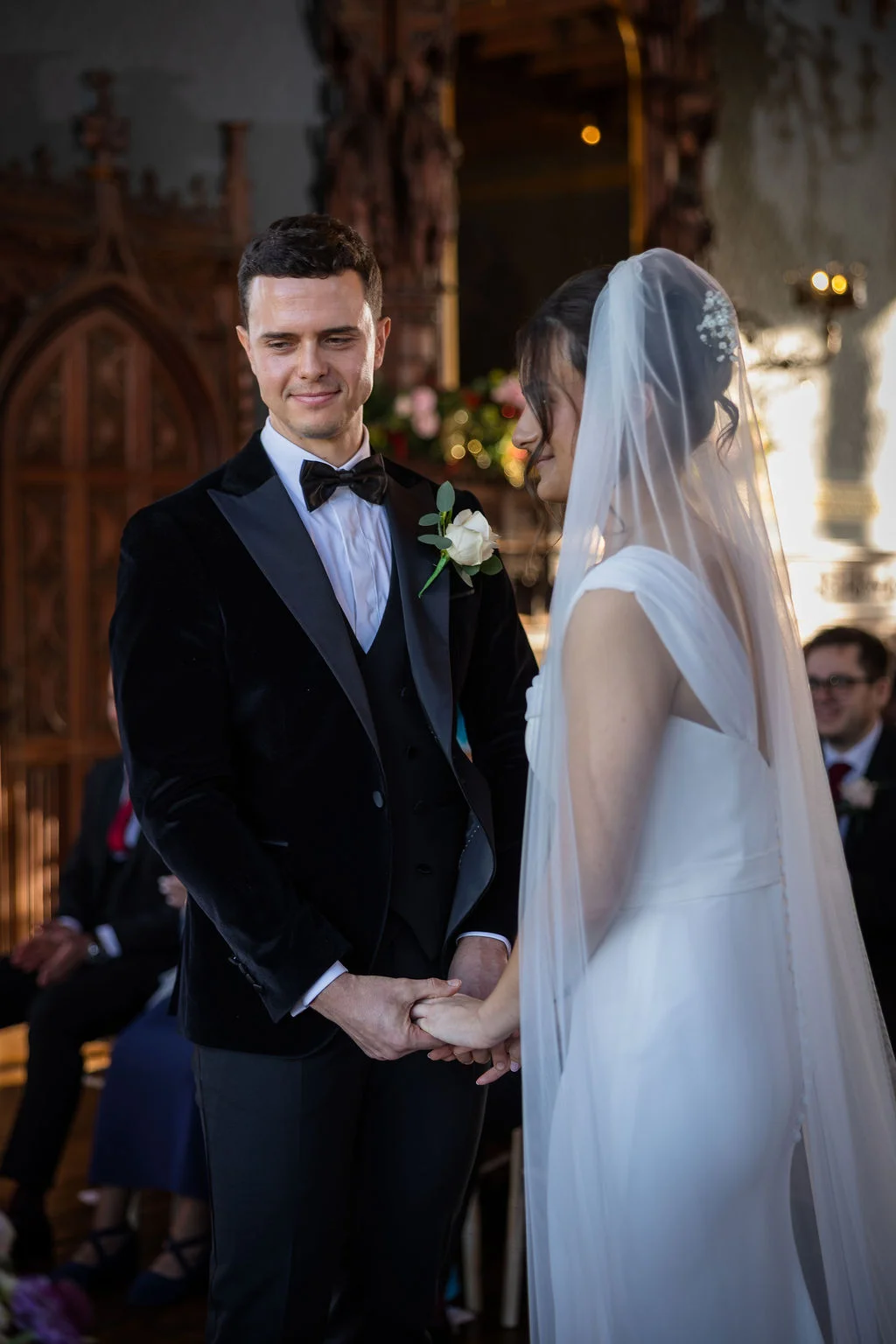 A groom in a black velvet tuxedo smiles warmly as he holds hands with his bride wearing a classic gown and veil inside a traditional church ceremony.