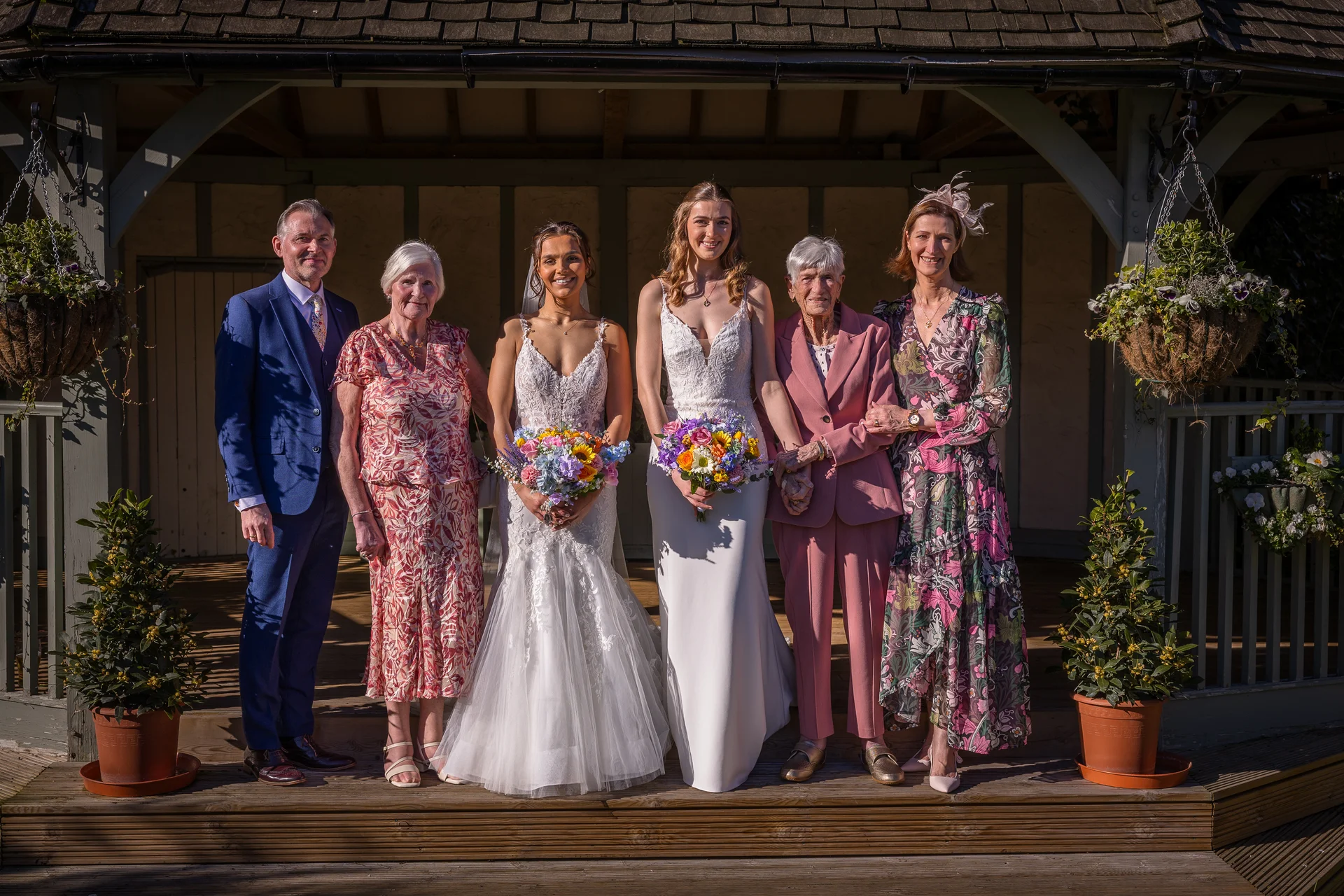 A smiling wedding party including two brides, family members, and older relatives pose together outside in the sunshine under a wooden gazebo.
