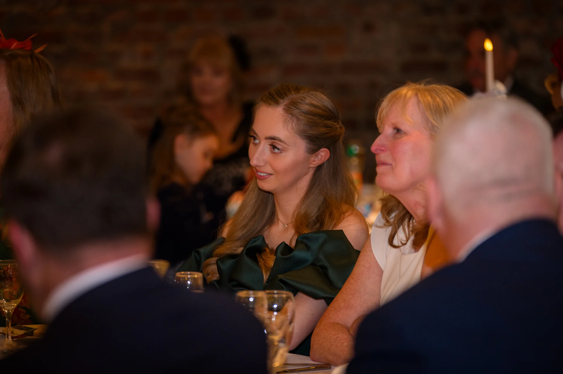 Wedding guests enjoy the wedding breakfast seated at tables in a rustic room with exposed brick walls and warm candle lighting.