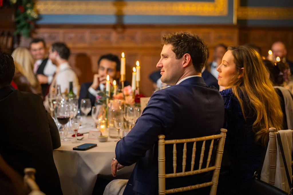 Wedding guests seated around a candlelit round table during the wedding breakfast in a wood-panelled reception room.