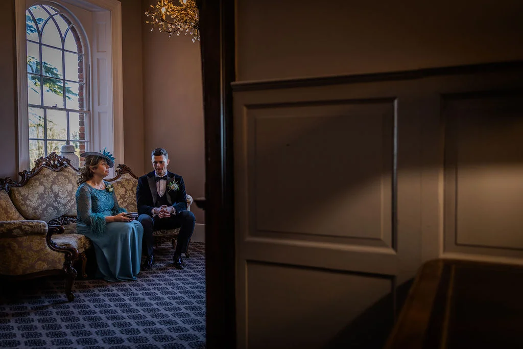 A groom in a black tuxedo and a mother of the bride in a blue dress wait quietly inside a panelled room with a large arched window.