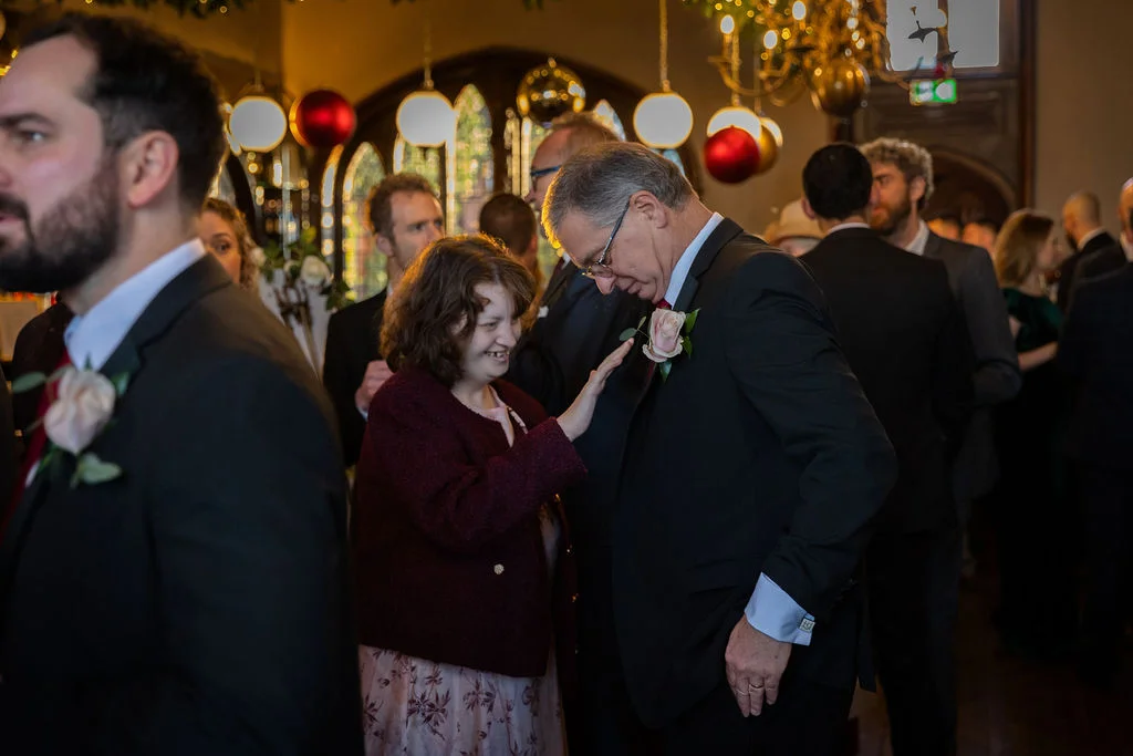 A guest smiling warmly and adjusting a boutonniere on a man wearing a black suit during a festive indoor wedding reception.