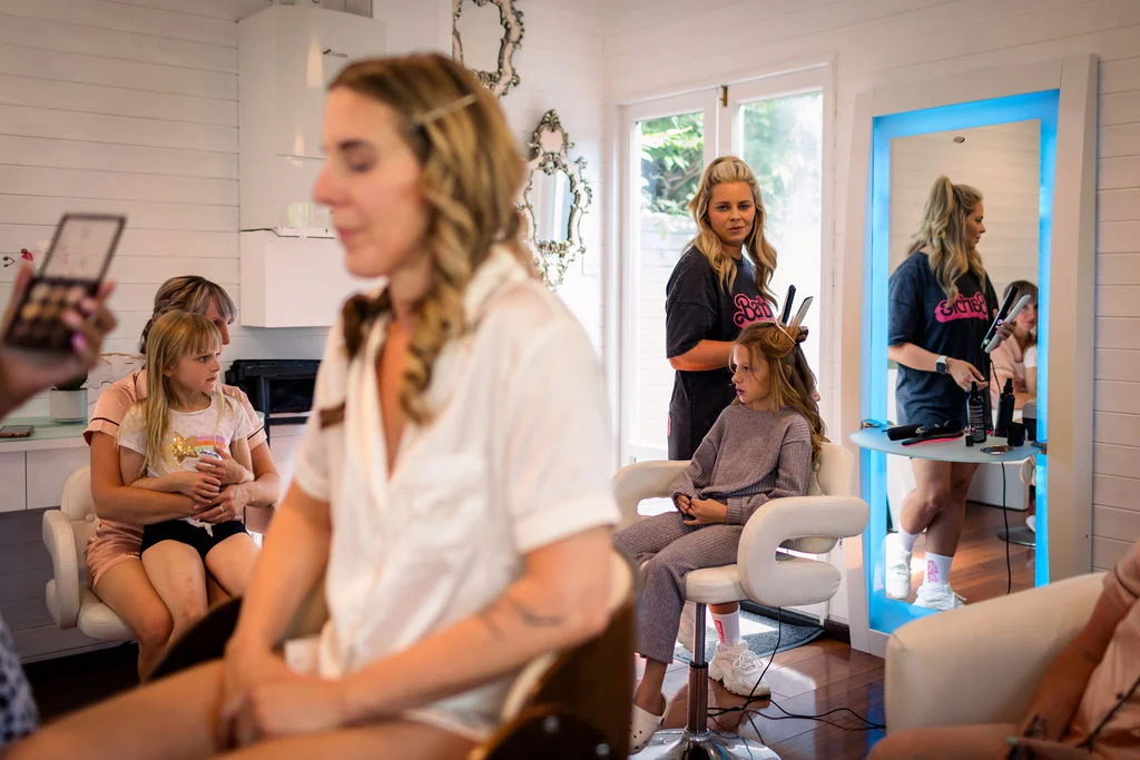 A bride sits in the foreground during morning preparations while a young flower girl and others have their hair styled inside a bright dressing room.