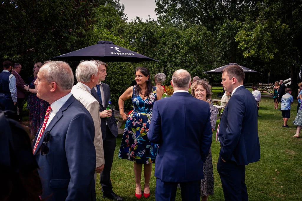 Wedding guests mingle and chat outdoors on a grassy lawn during a relaxed daytime drinks reception.