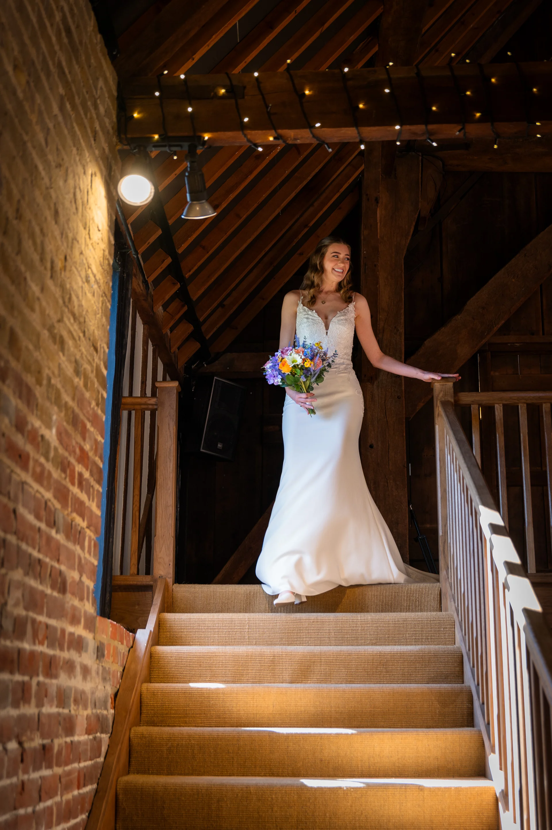 A smiling bride holding a colourful wildflower bouquet descends a carpeted wooden staircase inside a rustic oak framed barn adorned with fairy lights.
