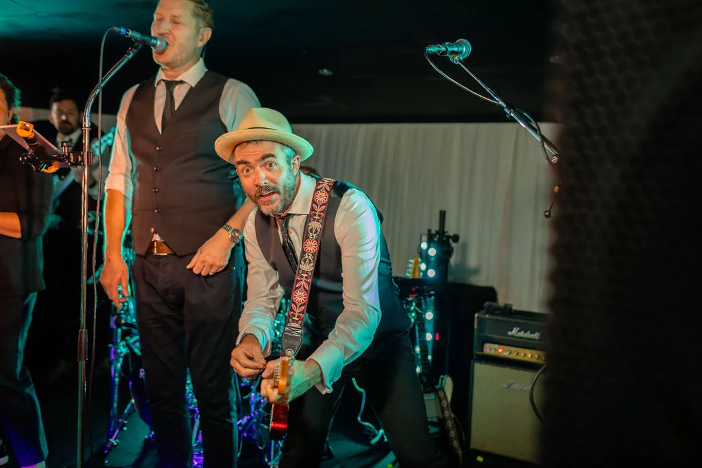 An energetic live wedding band performs on stage with a singer in a fedora playing guitar under colourful stage lighting.