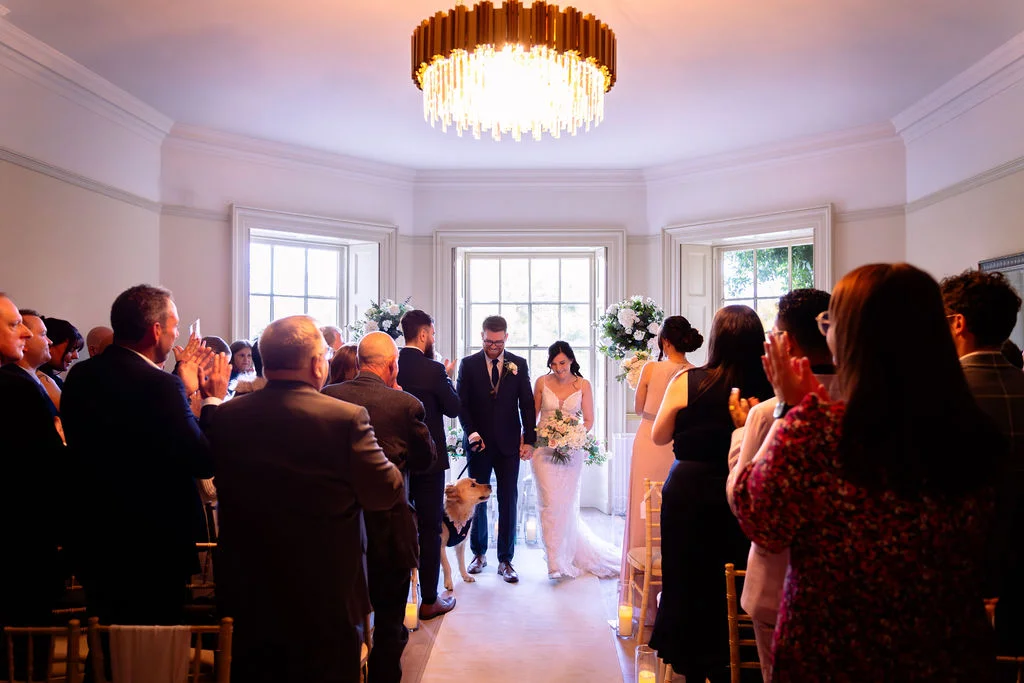 Newlyweds walk down the aisle together accompanied by their dog while guests applaud and celebrate during a bright indoor ceremony.