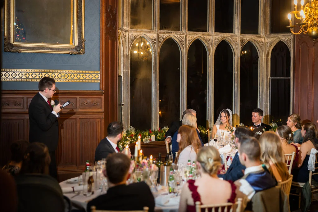 A guest delivers a speech at the top table during an elegant wedding reception inside a historic wood-panelled hall with arched windows.