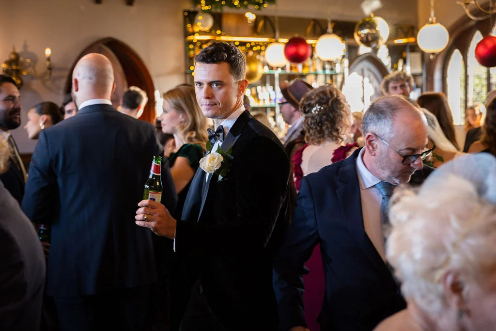 A stylishly dressed groom wearing a black tuxedo and holding a bottled drink glances over his shoulder amidst a lively crowd of wedding guests at an evening reception.