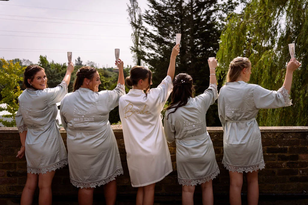 The bride and her four bridesmaids stand outside facing away from the camera, wearing matching robes and raising champagne flutes in a toast.