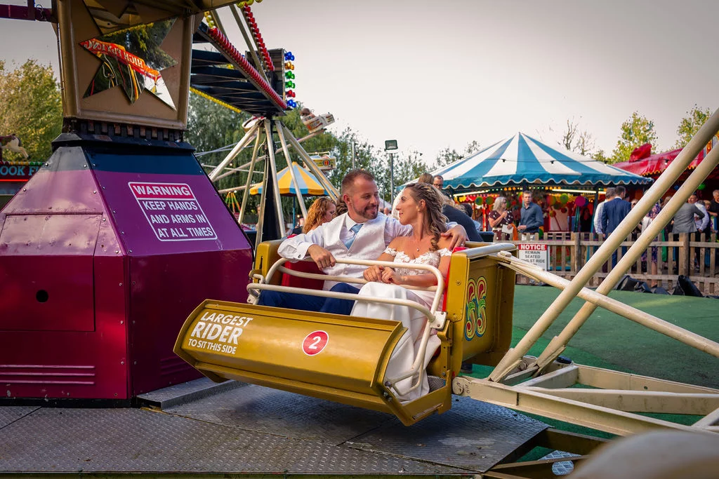 Newlyweds share a romantic moment sitting together in a vintage funfair ride at an outdoor festival reception.