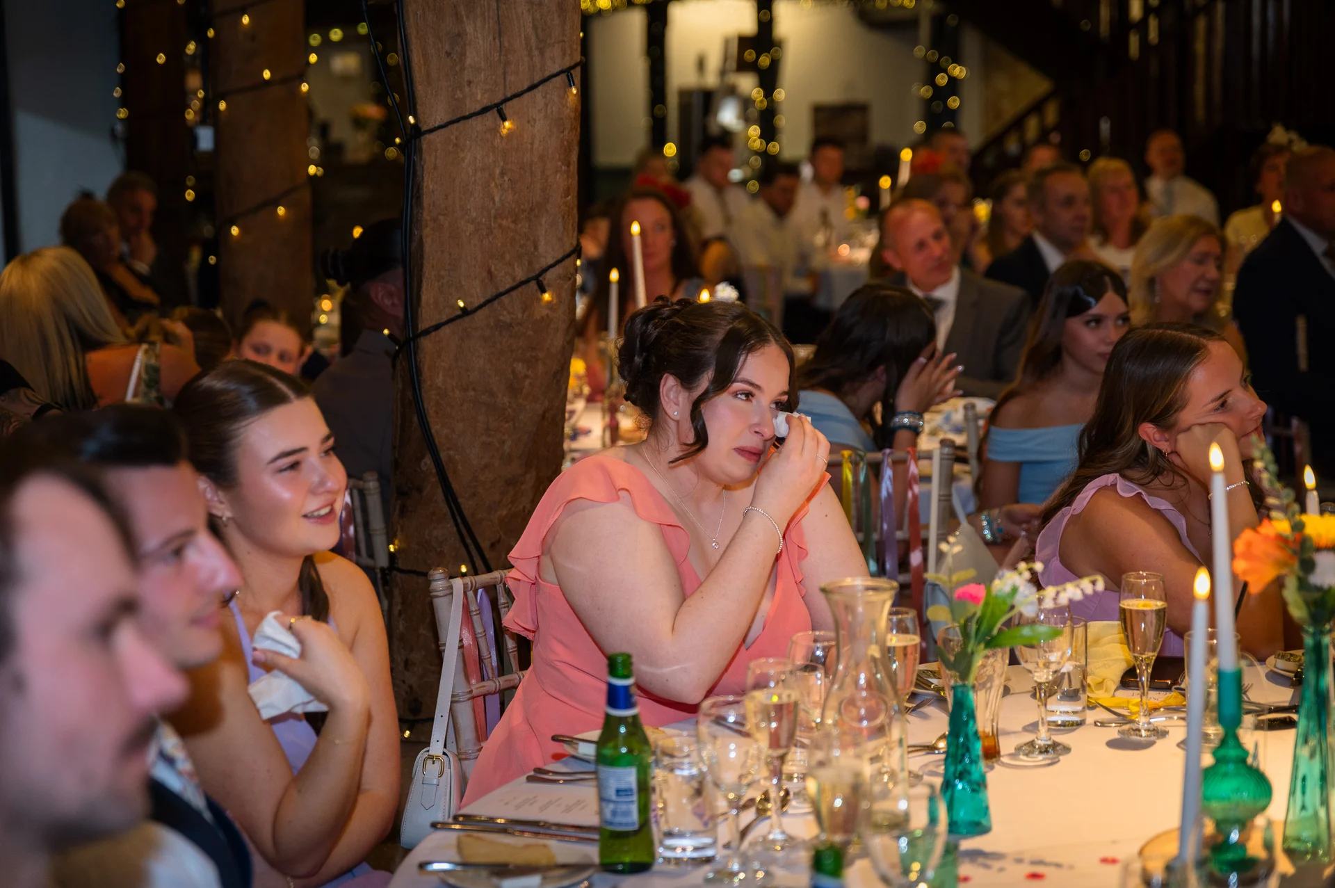 A guest at a wedding reception emotionally wipes a tear away during speeches inside a rustic barn illuminated by fairy lights and candlelight.