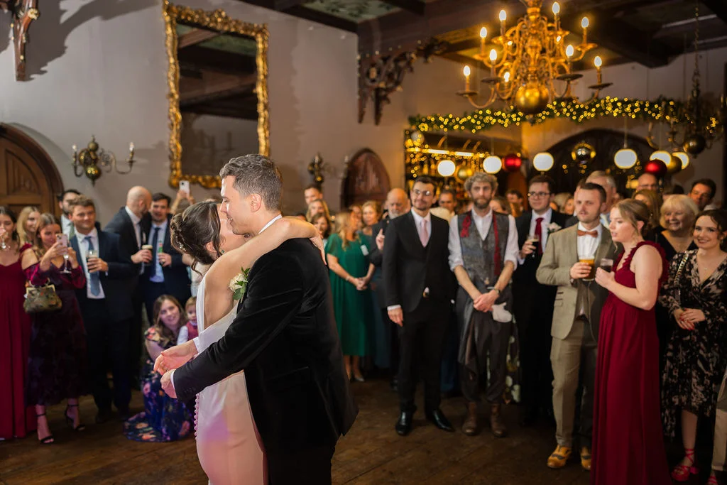 Newlyweds share an intimate embrace during their first dance inside a grand stately home ballroom adorned with chandeliers and warm fairy lights, surrounded by cheering guests.