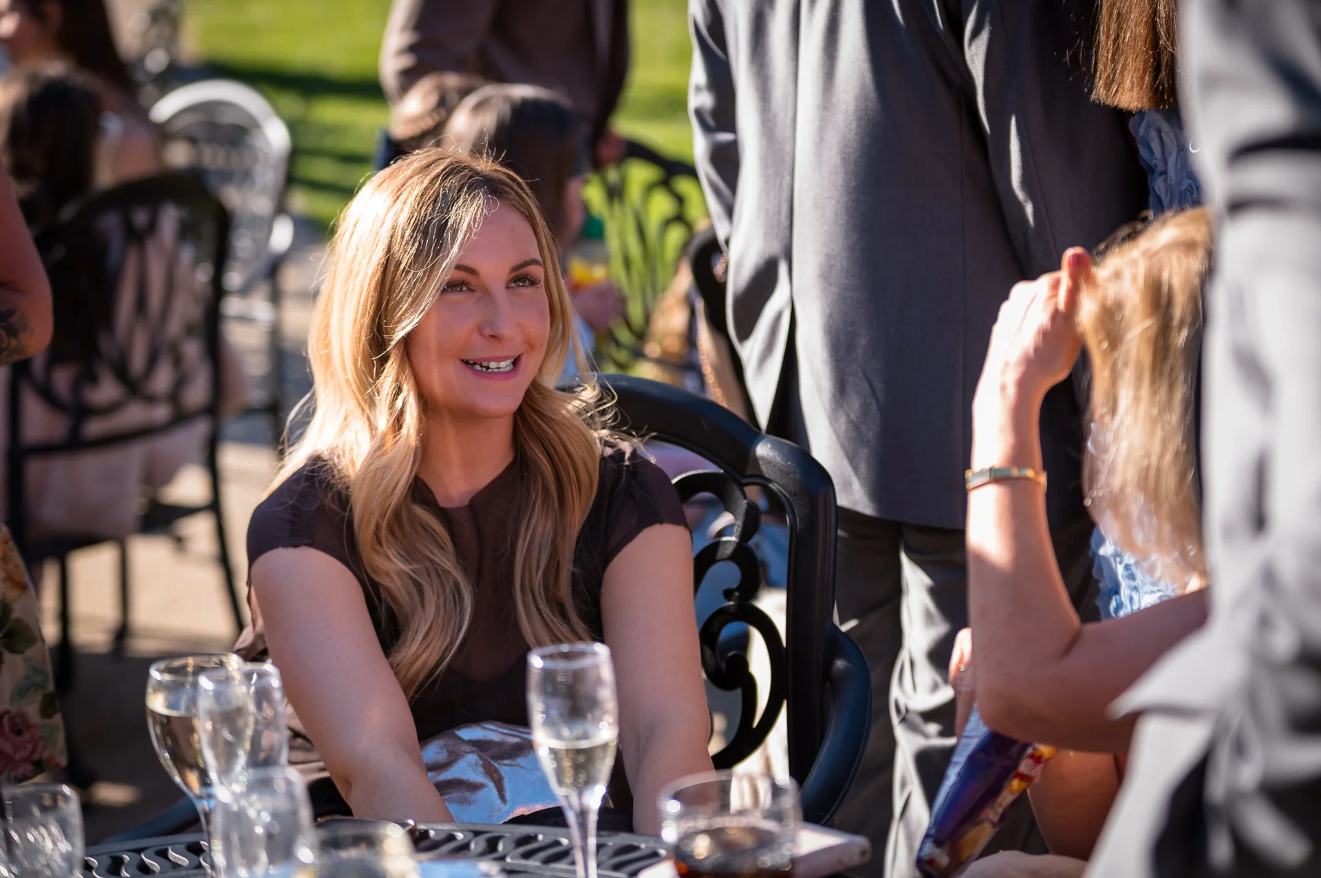 A blonde guest smiles warmly while seated outdoors at a wedding reception table surrounded by drinks and chatting guests.