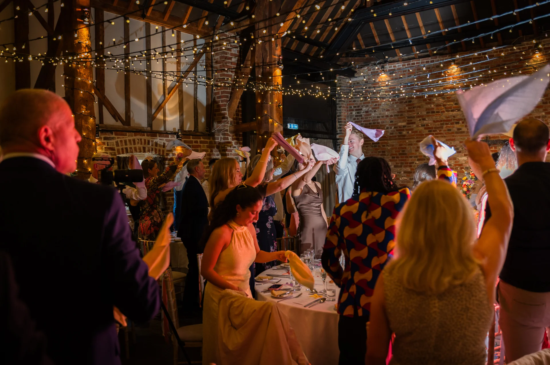 Wedding guests enthusiastically wave napkins in the air during the wedding speeches inside a rustic brick barn adorned with fairy lights.