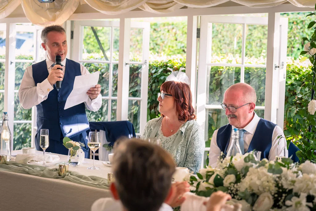 A man in a blue waistcoat delivers a speech at the top table during a wedding reception in a bright conservatory venue.