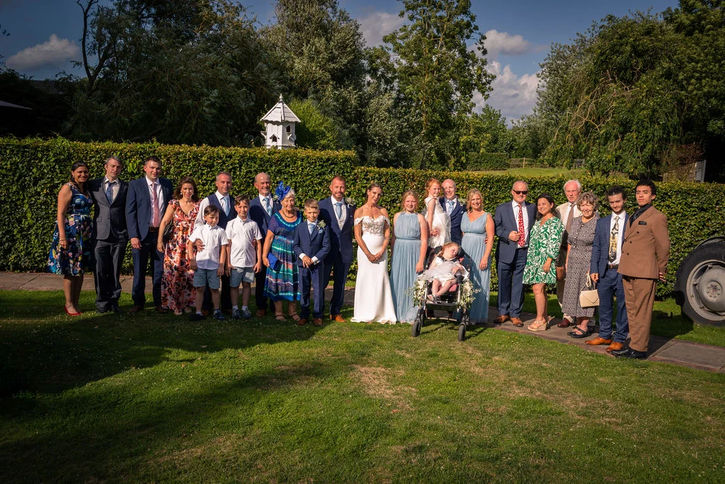 A large wedding group portrait stands outdoors on a lush green lawn in front of a neatly trimmed hedge under a bright blue summer sky.