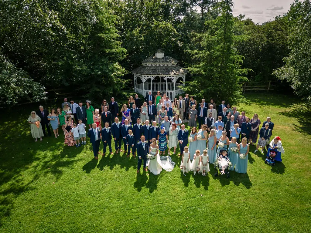 An aerial drone photograph captures a large wedding party and guests gathered on a manicured lawn in front of a white wooden gazebo on a sunny summer day.