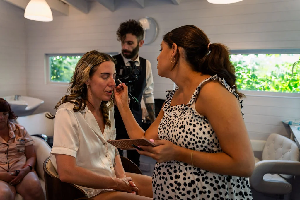 A bride sits in white silk pyjamas while a makeup artist applies cosmetics during morning preparations in a bright, modern room.