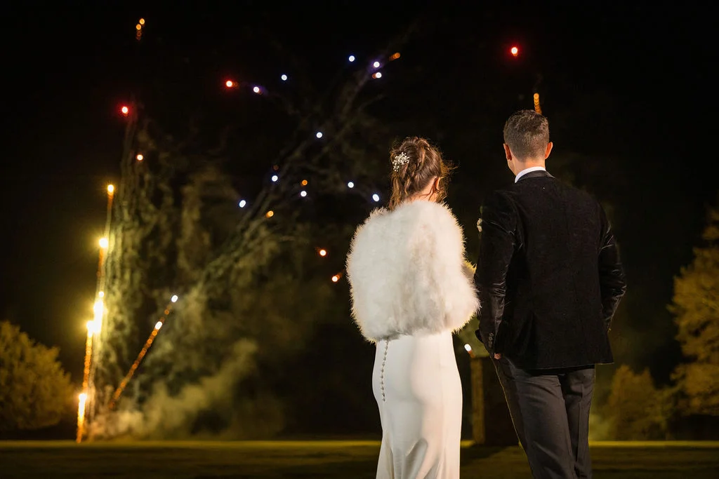 A newlywed couple watches a stunning nighttime fireworks display together outdoors, with the bride wearing a white faux fur stole and the groom in a dark jacket.