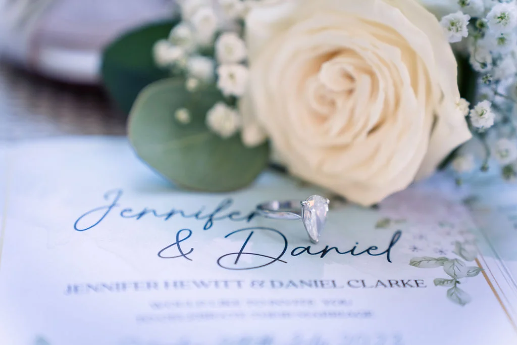 An engagement ring sits atop a personalised wedding invitation suite, accompanied by a white rose and delicate baby's breath florals.