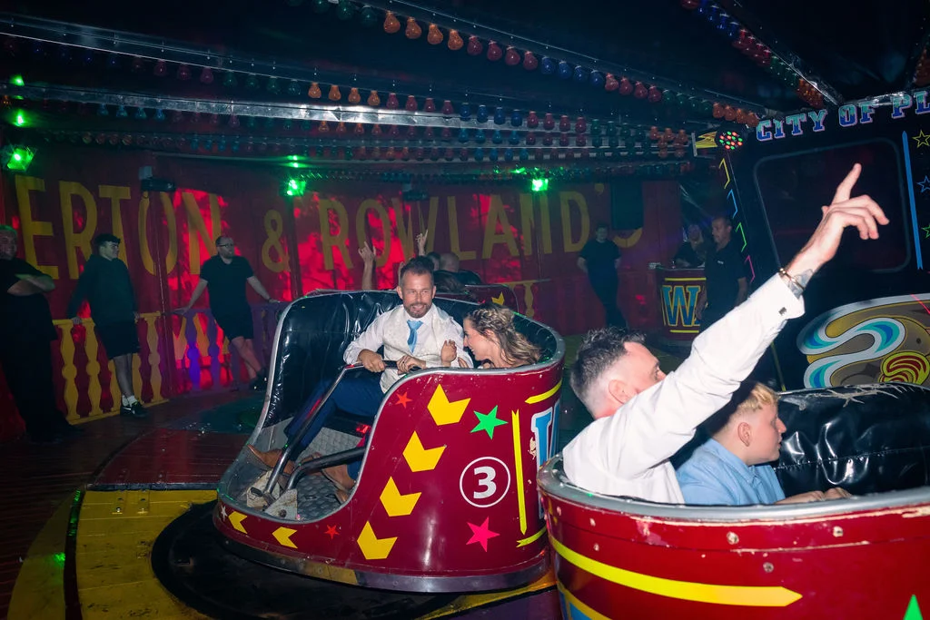Newlyweds and wedding guests laugh and enjoy a funfair ride together during an evening wedding reception.
