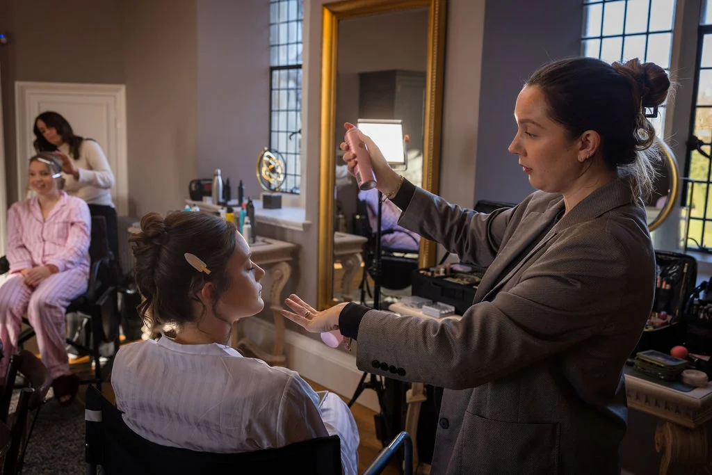 A professional hair and makeup artist sprays setting spray on a bride sitting in a makeup chair while bridesmaids relax in the background.