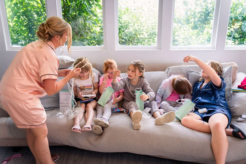 A woman wearing a pink satin robe stands by a sofa while opening gifts and sharing a morning moment with a group of children in matching pyjamas.