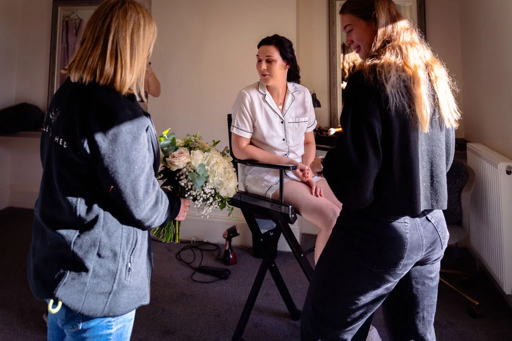 A bride sits in a makeup chair in white pyjamas while two women admire a floral bouquet during the morning wedding preparations.