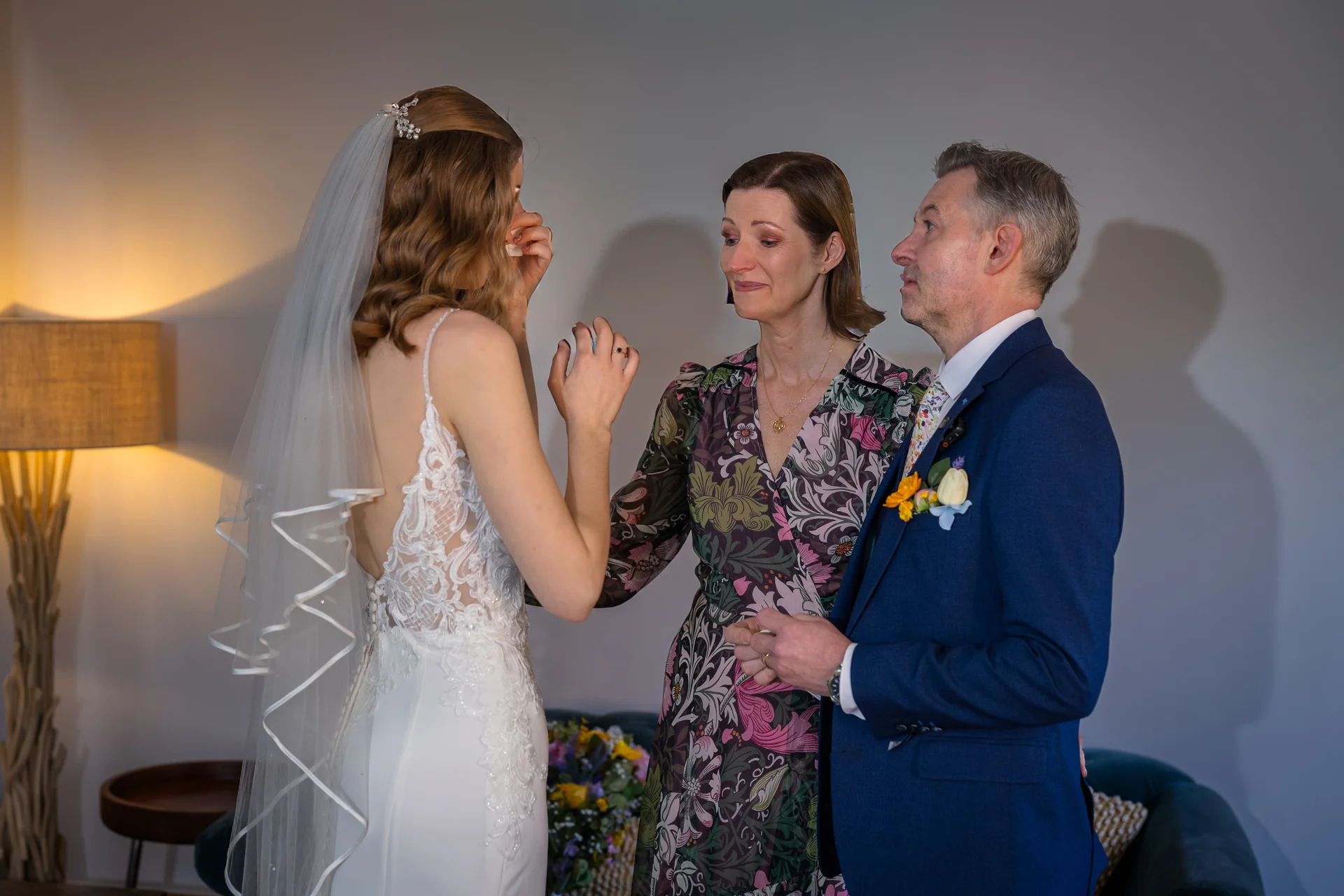 An emotional bride in a lace and crepe wedding gown wiping away happy tears as her mother and father look on tenderly indoors.