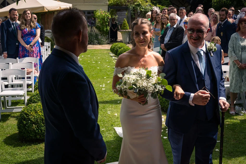 A smiling bride walks down a grassy outdoor aisle holding a white floral bouquet, linked with her father toward the waiting groom in a blue suit.