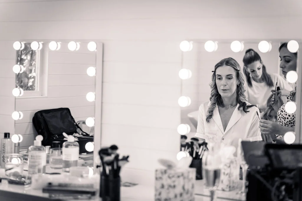 A bride and her bridesmaids getting ready in front of illuminated Hollywood style vanity mirrors during morning bridal preparations.